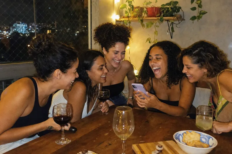 Amigas reunidas à mesa à noite, rindo, enquanto uma delas mostra uma conversa no celular